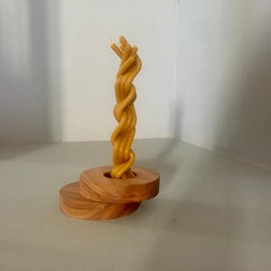 May include: A decorative candle holder featuring a twisted, light-yellow candle. The candle sits atop a wooden base composed of two circular tiers. The wood has a warm, natural tone, creating a contrast with the candle's color.