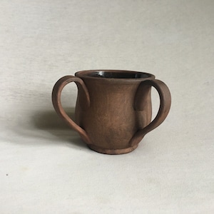 May include: A brown ceramic mug with two handles. The mug has a dark brown glaze and a slightly rounded shape.