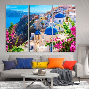 May include: A three-panel canvas print featuring a picturesque view of a white-washed village with blue-domed churches in Santorini, Greece. The image includes a vibrant blue ocean, a rocky cliffside, and lush green foliage with pink flowers.