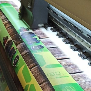 May include: A large format printer printing a green and brown design on a roll of material. The design features a wooden background with a green ski or snowboard.