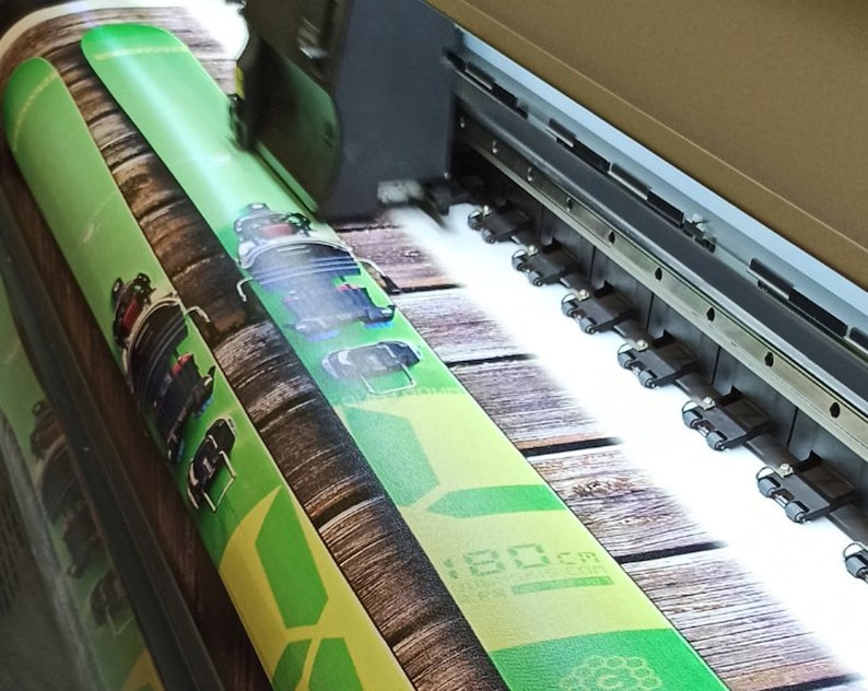 May include: A large format printer printing a green and brown design on a roll of material. The design features a wooden background with a green ski or snowboard.