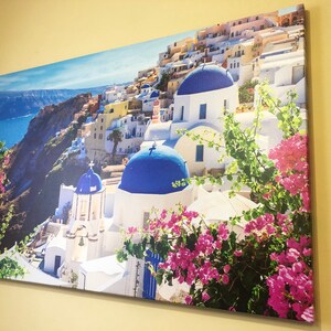 May include: A colorful canvas print featuring a picturesque view of a whitewashed village with blue-domed churches, nestled on a cliffside overlooking a sparkling blue sea. Lush green foliage and vibrant pink flowers add a touch of vibrancy to the scene.