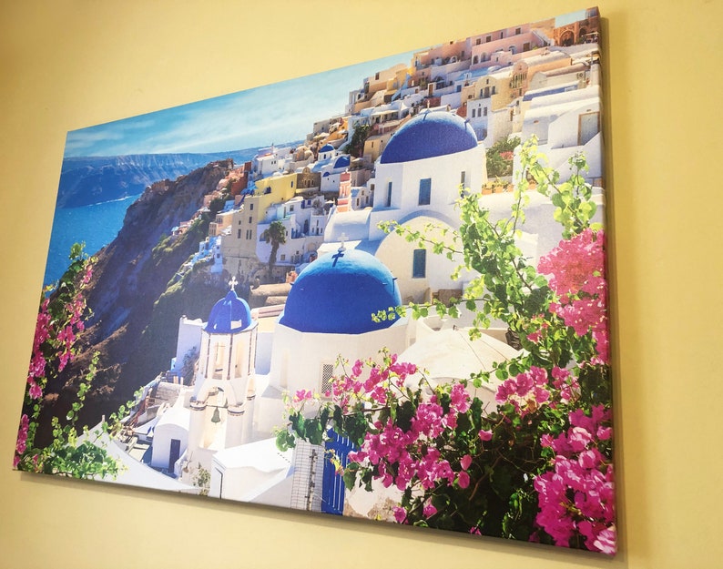 May include: A colorful canvas print featuring a picturesque view of a whitewashed village with blue-domed churches, nestled on a cliffside overlooking a sparkling blue sea. Lush green foliage and vibrant pink flowers add a touch of vibrancy to the scene.