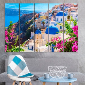 May include: A multi-panel canvas print featuring a picturesque view of white buildings with blue domed roofs in a Greek village. Lush green foliage with pink flowers frames the scene, creating a vibrant and colorful image.