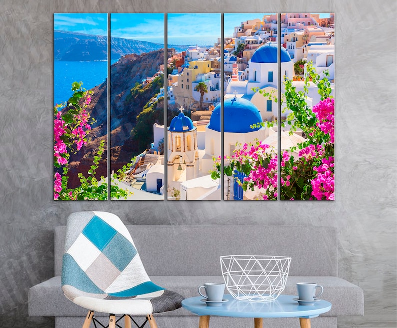 May include: A multi-panel canvas print featuring a picturesque view of white buildings with blue domed roofs in a Greek village. Lush green foliage with pink flowers frames the scene, creating a vibrant and colorful image.