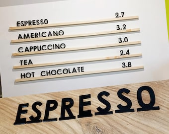 May include: A white sign with a wooden frame lists the prices of various coffee and tea drinks. The prices are listed in dollars. The sign reads: Espresso 2.7, Americano 3.2, Cappuccino 3.0, Tea 2.4, Hot Chocolate 3.8. Below the sign is a large black sign that reads "ESPRESSO".