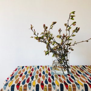 May include: A white tablecloth with a repeating pattern of colorful fish. A glass vase with a branch of green leaves sits on the tablecloth.