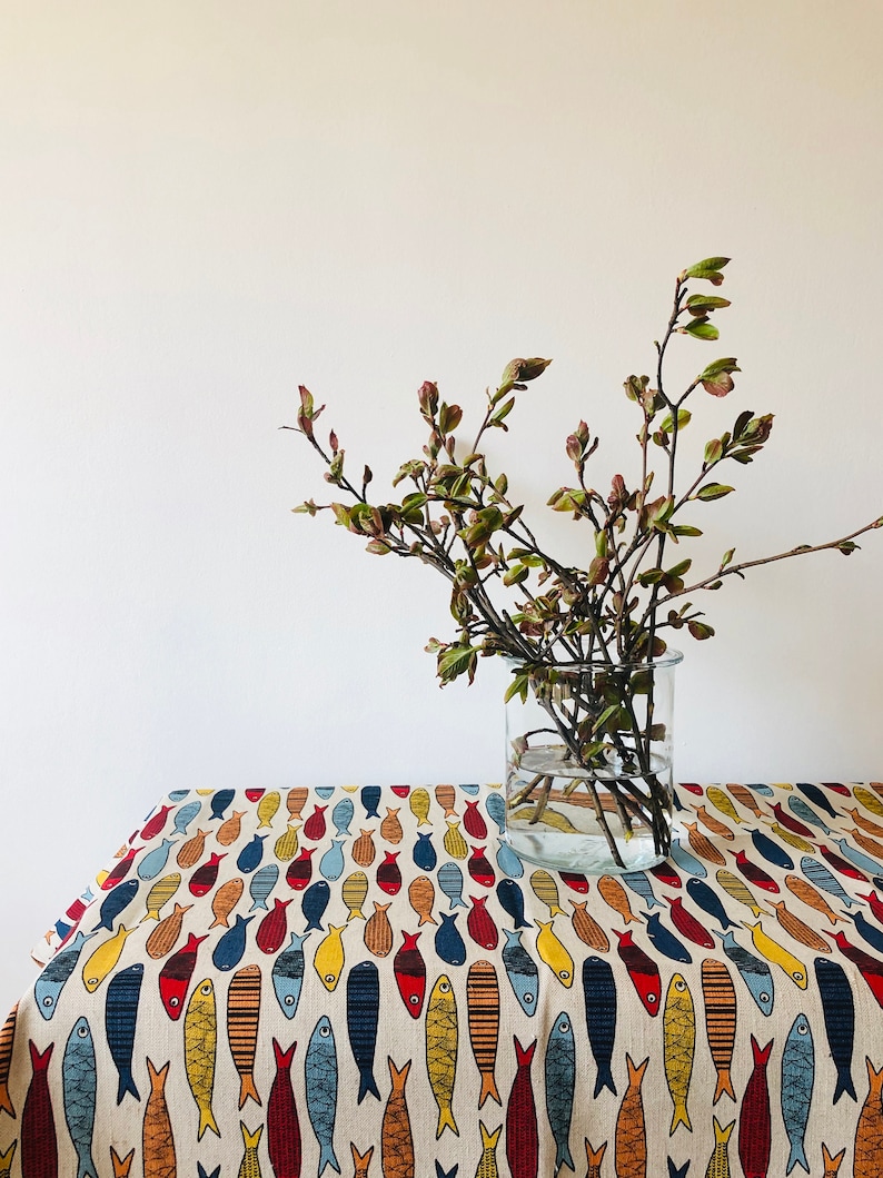 May include: A white tablecloth with a repeating pattern of colorful fish. A glass vase with a branch of green leaves sits on the tablecloth.