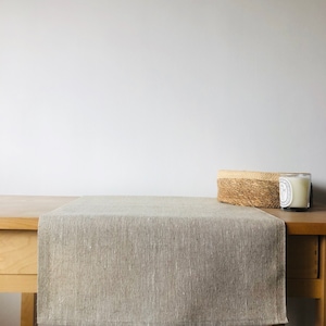 May include: A light brown linen table runner on a wooden table. The table runner is textured and has a subtle sheen.