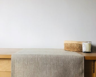 Puede incluir: Un camino de mesa de lino marrón claro sobre una mesa de madera. El camino de mesa es texturizado y tiene un brillo sutil.