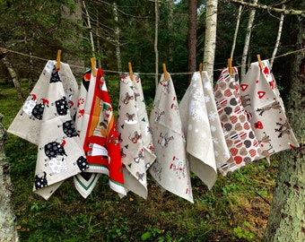 May include: Six different kitchen towels hanging on a clothesline in a woodland setting. The towels are made of linen and feature various festive designs, including reindeer, snowflakes, and mice.