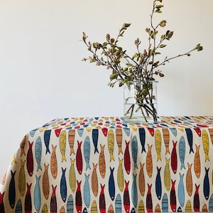 May include: A white tablecloth with a repeating pattern of colorful fish. The tablecloth is on a table with a vase of green branches in the center.