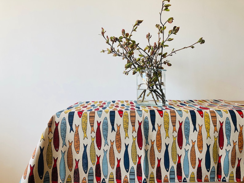 May include: A white tablecloth with a repeating pattern of colorful fish. The tablecloth is on a table with a vase of green branches in the center.