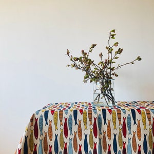 May include: A white tablecloth with a repeating pattern of colorful fish. The tablecloth is on a table with a vase of green branches in the background.