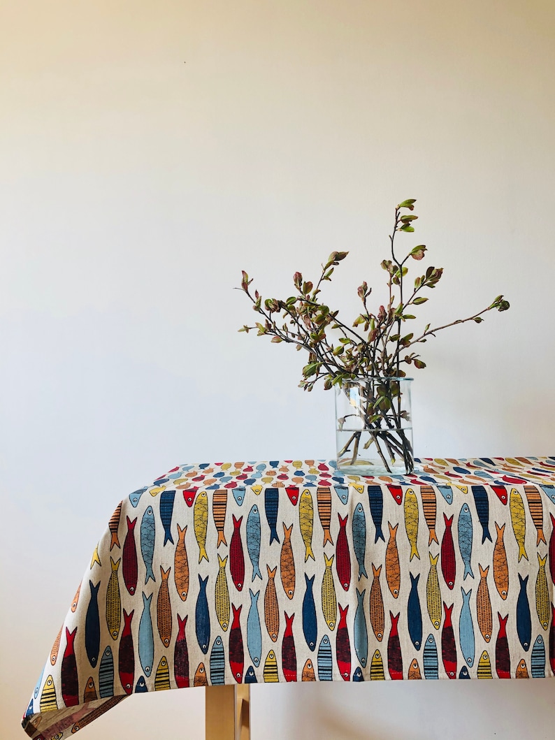 May include: A white tablecloth with a repeating pattern of colorful fish. The tablecloth is on a table with a vase of green branches in the background.