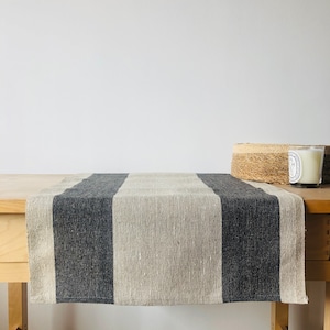 Könnte beinhalten: Ein gestreifter Tischläufer aus Leinen in den Farben Grau und Beige. Der Läufer liegt auf einem Holztisch.
