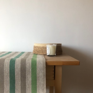 Puede incluir: Un camino de mesa de lino a rayas verdes y marrones sobre una mesa de madera con una cesta redonda tejida y una vela blanca en un tarro de vidrio.