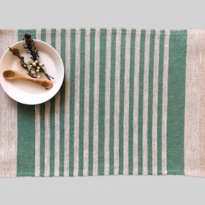 May include: A green and natural linen placemat with a white plate and wooden spoon on top. The placemat has a striped pattern.