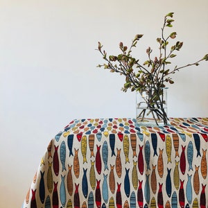 May include: A beige tablecloth with a repeating pattern of colorful fish. The tablecloth is draped over a wooden table with a vase of branches in the background.
