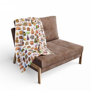 Puede incluir: Un sofá marrón con una manta estampada. La manta presenta ilustraciones coloridas de varios alimentos e insectos. El sofá tiene un asiento y respaldo rectangulares, con patas de madera. La manta está sobre el sofá.
