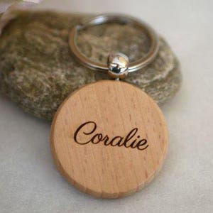 May include: A wooden keychain with the name "Coralie" engraved on it. The keychain is attached to a silver metal ring.