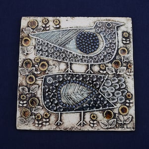 Pode incluir: Um azulejo de parede de cerâmica com fundo branco e dois pássaros estilizados em preto, branco e azul. Os pássaros são rodeados por desenhos florais em amarelo e castanho. O azulejo é assinado "LISA L."