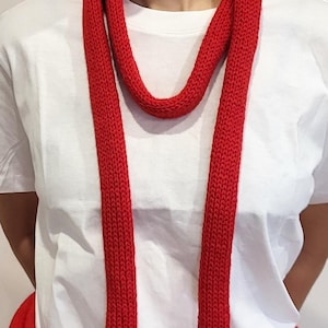 Può includere: Una sciarpa rossa brillante, lavorata a maglia, con una trama spessa e visibile, drappeggiata attorno al collo. Le lunghe estremità della sciarpa pendono dritte. La persona indossa una maglietta bianca. Un capo rosso abbinato è visibile in vita.