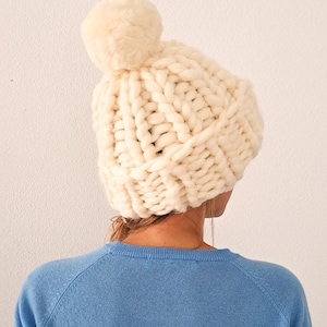 May include: A chunky knit beanie in an off-white cream colour, featuring a large, thick yarn texture and a matching pom-pom on top. The hat is constructed with a prominent cable-knit pattern and sits atop the head, providing full coverage.