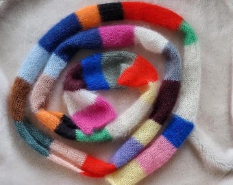 May include: A multi-coloured, hand-knitted scarf with a striped design. The scarf displays a range of colours, such as red, orange, blue, pink, and green. The scarf is made from a soft, fuzzy yarn and is coiled on a light-coloured surface.