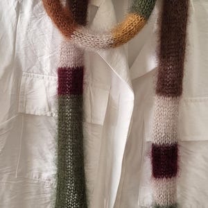 Könnte beinhalten: Ein farbenfroher, handgestrickter Schal mit weicher, flauschiger Textur. Der Schal hat eine Mischung aus Braun-, Grün- und Cremetönen mit burgunderfarbenen Streifen. Der Schal ist über einem weißen Hemd drapiert.