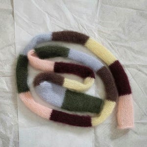 Könnte beinhalten: Ein mehrfarbiger Strickschal mit Streifenmuster. Der Schal hat Abschnitte in Rosa, Hellblau, Grün, Gelb, Braun und Burgunder. Der Schal ist aus einem weichen, flauschigen Material gefertigt und auf einer weißen Oberfläche aufgerollt.