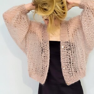 May include: A light pink, open-weave cardigan with long sleeves and two buttons. The cardigan is worn over a black dress. The sleeves are wide and the cardigan has a loose fit. The style is a light, airy, and feminine.