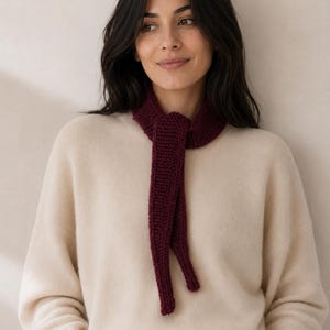 Pode incluir: Um cachecol de malha em tom bordô profundo com uma textura de ponto musgo, atado com um nó simples à volta do pescoço. O cachecol apresenta pontas longas e estreitas que caem, contrastando com uma camisola bege de manga comprida.