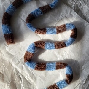 Könnte beinhalten: Ein gestrickter Schal mit abwechselnden Streifen in Hellblau und Braun. Der Schal ist aus einem weichen, strukturierten Garn gefertigt und wird auf einer weißen, zerknitterten Oberfläche präsentiert. Das Design des Schals ist ein einfaches, aber auffälliges Muster.