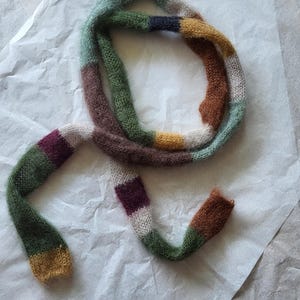 Könnte beinhalten: Handgestrickter Schal mit Streifenmuster in verschiedenen Farben wie Grün, Braun, Lila und Gelb. Der Schal besteht aus weichem, strukturiertem Garn und liegt auf einer weißen Oberfläche.