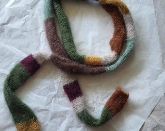 May include: A multi-coloured, hand-knitted scarf with a striped design. The scarf features shades of green, brown, purple, and yellow. It is made from a soft, textured yarn and is laid out on a white surface.