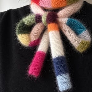 May include: A fuzzy, knitted scarf featuring a vibrant, multi-coloured striped pattern. The scarf is tied in a bow around the neck, displaying blocks of red, orange, olive green, light pink, dark purple, teal, white, navy blue, and light blue.