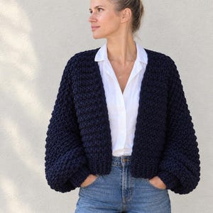 Könnte beinhalten: Eine dunkelblaue, grob gestrickte Strickjacke mit offener Vorderseite, langen Ballonärmeln und gerippten Bündchen. Das Kleidungsstück wird über einem weißen Hemd mit Kragen und einer blauen Jeans getragen und hat eine dicke, strukturierte Optik.