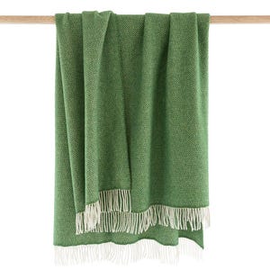 Puede incluir: Una manta de lana verde con diseño de espiga y flecos blancos. La manta está doblada por la mitad y colgada de una barra de madera.