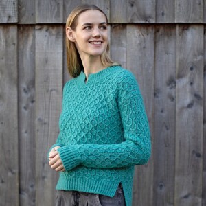 Puede incluir: Un suéter de punto azul turquesa con un patrón texturizado. El suéter tiene un cuello redondo y mangas largas.