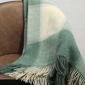 Puede incluir: Una manta a cuadros verde y crema con flecos. La manta está sobre una silla marrón. El patrón de cuadros presenta cuadrados de varios tonos de verde y crema. Los flecos son de color crema.