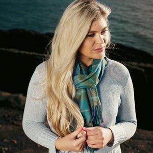 Könnte beinhalten: Eine Frau trägt einen hellgrauen Pullover und einen karierten Schal in Türkis und Grün mit Fransen. Der Schal hat Akzente in Orange und Lila. Die Frau hat lange blonde Haare und blickt nach unten.