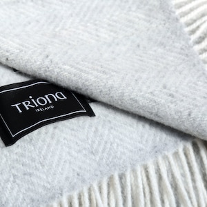 Puede incluir: Una suave manta de lana gris con flecos blancos. La manta está doblada por la mitad y tiene una etiqueta negra que dice "Triona Ireland".