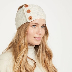 Puede incluir: Diadema de punto color crema con diseño de punto trenzado. La diadema presenta tres botones redondos de madera marrón en el lateral. El accesorio está diseñado para mantener las orejas calientes y es adecuado para climas fríos.