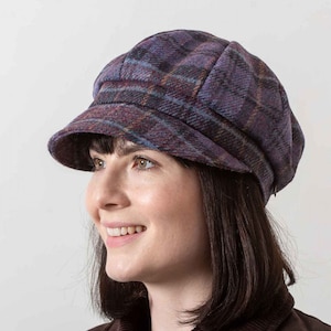 Puede incluir: Una gorra de estilo newsboy de cuadros azules y morados con un ala ancha. La gorra está hecha de un tejido suave y texturizado.