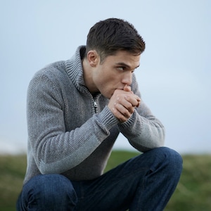 Puede incluir: Un hombre con un suéter gris y pantalones vaqueros azules se sienta con las manos juntas frente a él, mirando hacia un lado.