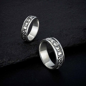 Puede incluir: Dos anillos de plata con un diseño de nudo celta y un símbolo Claddagh. Los anillos están sobre una superficie negra.