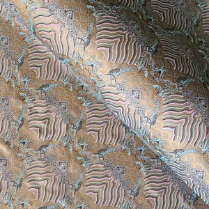 May include: Close-up of a fabric with a repeating pattern of stylized waves and cloud-like shapes. The design features a blend of gold, brown, light blue, and pink hues, creating a textured and layered appearance. The fabric has a slight sheen.