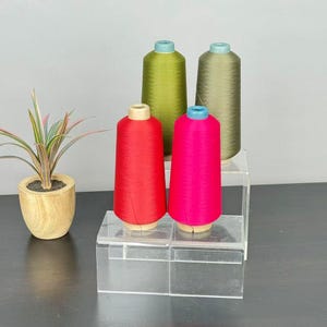 May include: Four spools of thread arranged on clear acrylic risers. Two spools in the front are bright red and vibrant pink, while two in the back are olive green and muted sage. A small potted plant with variegated leaves sits to the left.