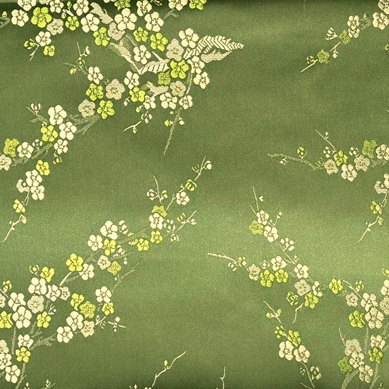 May include: Green fabric with a repeating pattern of white and yellow floral branches. The flowers have a detailed, textured appearance, and the background is a solid, muted green.